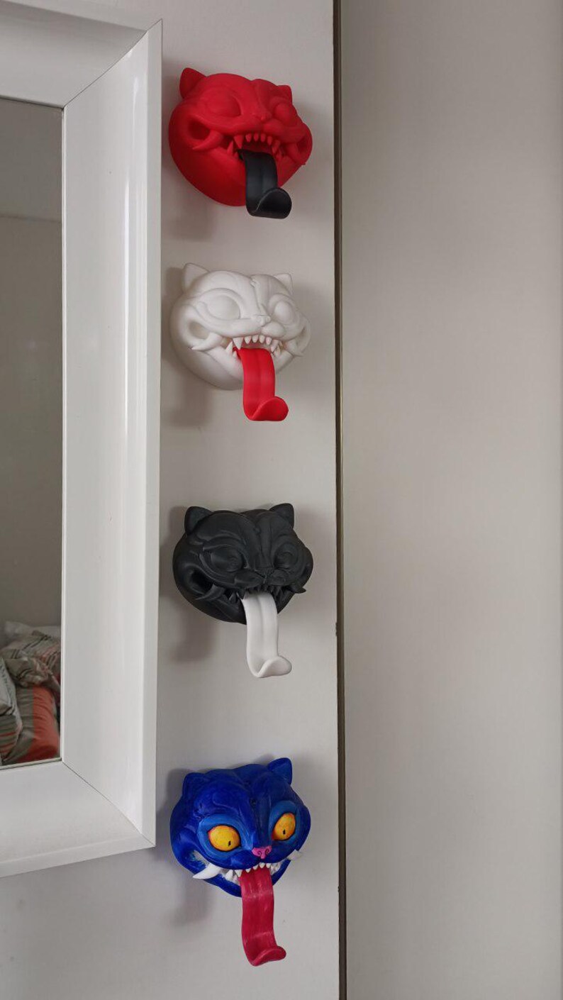 May include: Four cat-shaped wall hooks in red, white, black, and blue. Each hook has a protruding tongue that serves as the hook. The cat faces have sharp teeth and are mounted on a white wall.