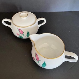 May include: A white ceramic sugar bowl with a lid and a creamer. Both pieces have a gold trim and are decorated with pink rose buds and green leaves. The sugar bowl has two handles, and the creamer has one handle and a spout.