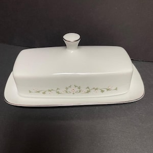 May include: White ceramic butter dish with a lid and a decorative floral design. The dish has a silver trim and a small handle on the lid. The floral design is in shades of green and pink.
