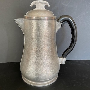 May include: A vintage silver-toned coffee pot with a textured surface and a black handle. The pot has a lid with a decorative handle and a spout for pouring. The design is classic and functional.