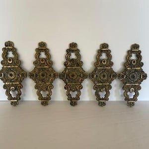 May include: Five ornate gold-toned metal drawer pulls with intricate designs. Each pull features a central diamond shape surrounded by swirling patterns.