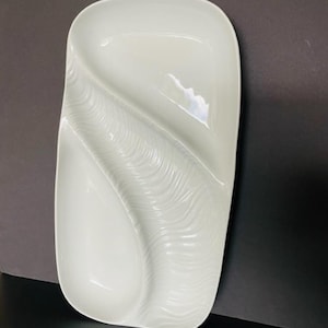 May include: A white, rectangular serving platter with a unique, wave-like design. The platter features a central, textured ridge that divides the surface into two sections. The overall shape is elongated with rounded corners, suitable for serving appetizers or small dishes.