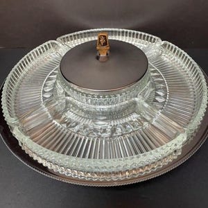 May include: A circular clear glass divided serving platter featuring a central lidded compartment with a dark metallic lid and a small brown decorative finial. The glass tray has a radial ribbed pattern and a textured diamond-cut outer edge.