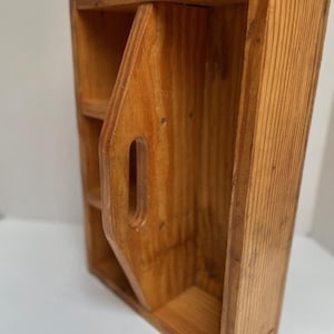 May include: A wooden storage box with a handle and multiple compartments. The box is made of light brown wood and has a rustic appearance. The handle is cut out of the wood, and the box appears to be handmade.