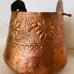 May include: A copper bucket with a floral design. The bucket has a handle on the side and a decorative lip.