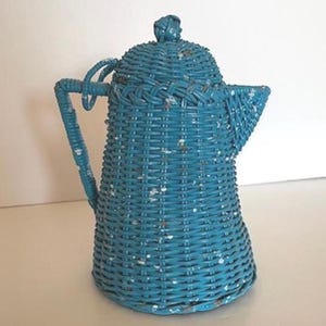 May include: A turquoise wicker teapot with a lid and handle. The teapot has a woven design and is painted with a distressed finish, showing white speckles. The spout is triangular, and the handle is curved.