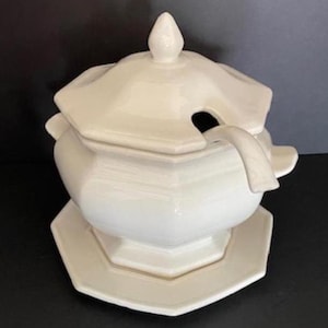 May include: A white ceramic soup tureen with a lid and a matching saucer. The tureen has a geometric design and a handle on the side. The lid has a small knob on top and a slot for a ladle.