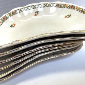 May include: A stack of six off-white, scalloped-edge serving dishes. Each dish has a gold rim and a decorative border with a green lattice pattern and small floral accents in orange and yellow. The dishes are stacked at an angle.