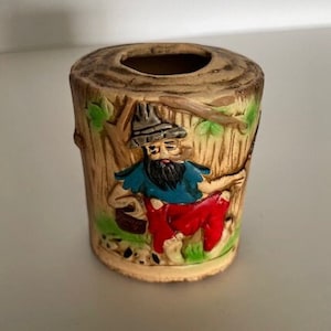 May include: A ceramic planter shaped like a tree stump with a bearded man in a blue shirt and red pants sitting on the stump. The man is holding a stick and there are two small dogs at his feet.