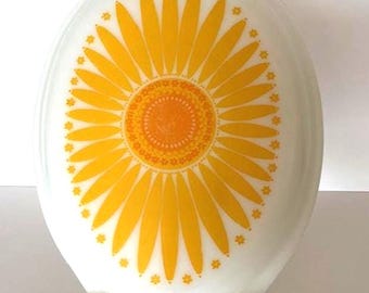 May include: White oval casserole dish with a large yellow sunflower design. The sunflower has a bright orange center and yellow petals. The dish is on a gold-colored metal stand.