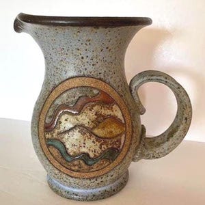 May include: A speckled ceramic pitcher with a dark brown rim and handle. The pitcher features a circular design with abstract landscape elements in shades of brown, red, and teal. The pitcher has a wide opening and a sturdy base.