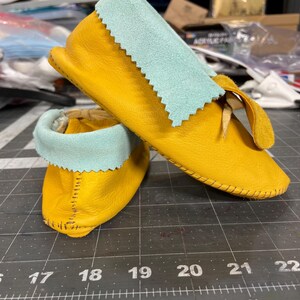 May include: A pair of yellow leather moccasins with light blue suede cuffs featuring a pinked edge. The moccasins have a visible whipstitch detail along the heel and are positioned on a black cutting mat with white measurement markings in inches.