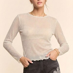Könnte beinhalten: Ein durchsichtiges Langarm-Top mit schimmerndem, glitzerndem Aussehen. Das Top hat einen runden Ausschnitt und gerüschte Manschetten. Das Model trägt eine schwarze Jeans mit Kreuzverzierungen auf den Vordertaschen.