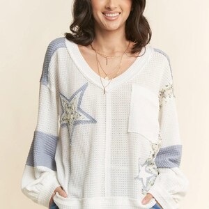 May include: White waffle-knit long-sleeve top with a V-neck, featuring light blue accents on the sleeves and star appliqués. The top includes a chest pocket and floral detailing, creating a casual yet stylish look.