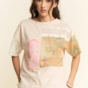 以下が含まれることがあります： クリーム色のパッチワークデザインのTシャツ。ピンクの抽象的な形、花柄、袖と肩のレースのディテールが特徴です。カジュアルウェアに最適な、スタイリッシュでユニークなトップスです。