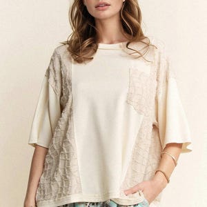 Può includere: Top color crema a maniche corte con pannelli strutturati e una tasca. Il top ha una vestibilità ampia ed è abbinato a pantaloni fantasia. Braccialetti dorati adornano il polso.