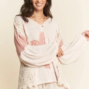May include: Cream waffle-knit top with pink panels and lace inserts. The long-sleeved top has a V-neck and a loose fit. The sleeves are gathered at the cuffs, and the lace trim adds a delicate touch. The outfit is completed with matching shorts.