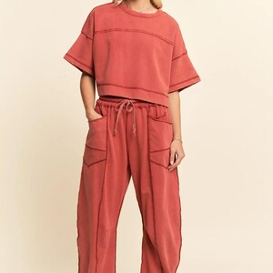 May include: A terracotta-colored two-piece lounge set. The short-sleeved top has a crew neck and horizontal seam details. The matching pants have a drawstring waist, pockets, and visible stitching. The outfit is made of a soft, comfortable material.