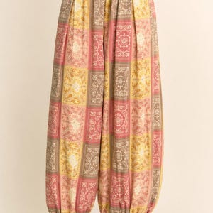 May include: Wide-leg trousers with a patchwork design in pink, yellow, and brown. The fabric features a repeating paisley-like pattern within each square. The trousers have a drawstring waist and are gathered at the ankles. Black heeled sandals complete the look.