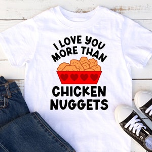 May include: White t-shirt with a black graphic that says "I love you more than chicken nuggets". The graphic features a red bowl with chicken nuggets and hearts.
