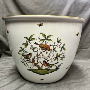 May include: A white ceramic planter with a gold rim, featuring a detailed illustration of birds perched on a tree with green leaves and butterflies. The planter has a wide, rounded shape, suitable for indoor or outdoor use.