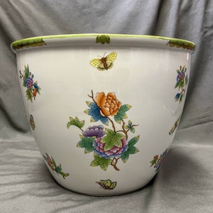 May include: A white ceramic planter with a green rim and floral and butterfly designs. The planter features a large orange and purple flower arrangement with blue and green leaves. Several butterflies are scattered around the planter.