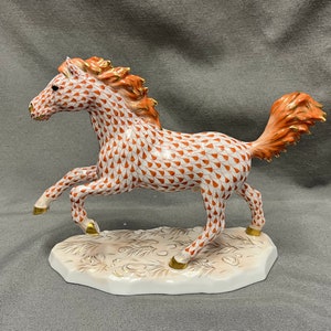 May include: A ceramic figurine of a running horse. The horse is white with a pattern of orange teardrops. The mane and tail are orange with gold accents. The hooves are gold, and the base is white with gold detailing.