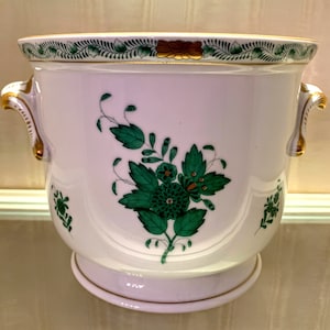 May include: White ceramic planter with green floral design and gold accents. The planter has two handles and a decorative rim. The floral design features a central flower with surrounding leaves. The planter is likely for indoor use.