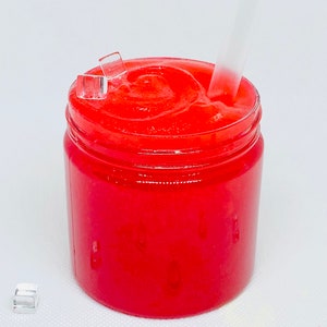 May include: A clear glass jar filled with a bright red, slushy-like substance. A clear plastic straw is sticking out of the top of the jar. There are small, clear plastic ice cubes scattered around the jar.