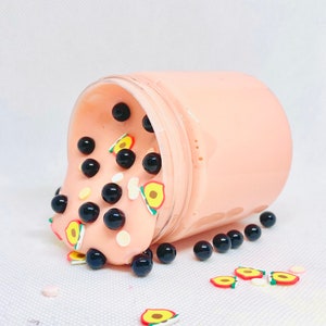 May include: A pink slime with black boba balls and peach slices inside a clear jar. The slime is spilling out of the jar onto a white surface.