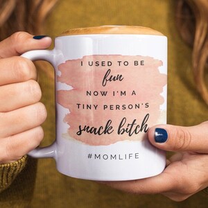 Pu&ograve; includere: Tazza in ceramica bianca con la scritta "I USED TO BE fun NOW I'M A TINY PERSON'S snack bitch #MOMLIFE" in nero e un design acquerello rosa. La tazza &egrave; piena di una bevanda ed &egrave; tenuta da una persona.
