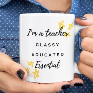 May include: White ceramic mug with the text "I'm a teacher, Classy, Educated, Essential" in black script. Yellow star accents are scattered around the text. The mug is held by a person wearing a blue button-down shirt.