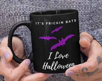 May include: Black ceramic mug with the text "IT'S FRICKIN BATS" and "I Love Halloween" in white. The mug features three purple bat illustrations. The mug is held by a person.