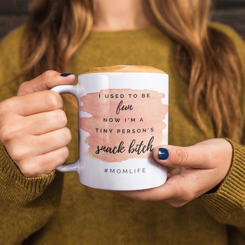 Pu&ograve; includere: Tazza in ceramica bianca con la scritta "I USED TO BE fun NOW I'M A TINY PERSON'S snack bitch #MOMLIFE" in nero e un design acquerello rosa. La tazza &egrave; piena di una bevanda ed &egrave; tenuta da una persona.