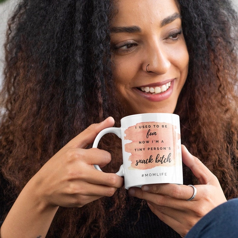 Pu&ograve; includere: Tazza in ceramica bianca con manico curvo, con la scritta "I USED TO BE fun NOW I'M A tiny person's snack bitch" in un mix di font e colori. La tazza ha uno sfondo color pesca in stile acquerello e l'hashtag #MOMLIFE.