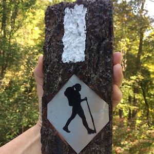 Könnte beinhalten: Ein Holzschild mit einem weißen Blitz und einem rautenförmigen Metallschild mit einer Wanderer-Silhouette. Der Wanderer trägt einen Rucksack und einen Wanderstock.