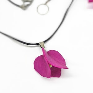 Könnte beinhalten: Eine Halskette mit einem Anhänger in Form einer leuchtend magentafarbenen Bougainvillea-Blüte, der an einem dünnen schwarzen Band hängt. Der Anhänger ist mit einer silberfarbenen Metallöse befestigt. Ein passendes magentafarbenes Blütenblatt ist oben rechts zu sehen.