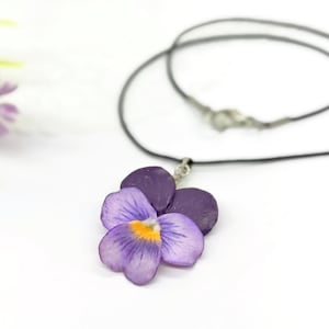 Könnte beinhalten: Eine Halskette mit einem Anhänger in Form eines violetten Stiefmütterchens mit gelber Mitte. Der Anhänger ist an einem dünnen schwarzen Band mit einem silberfarbenen Metallverschluss befestigt. Die Blütenblätter zeigen helle und dunkle Violetttöne.