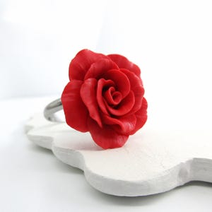May include: A red rose ring with a detailed floral design. The ring features a large, vibrant red rose as the focal point, set on a silver-colored band. The rose is intricately sculpted, showcasing the delicate petals. The ring is displayed on a white, decorative surface.