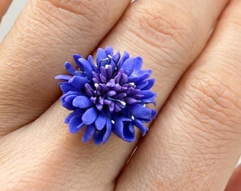 May include: A ring featuring a vibrant blue flower centerpiece with multiple layered petals and a detailed, textured center. The floral design is circular and sits prominently on the finger, showcasing a deep, saturated blue color throughout.