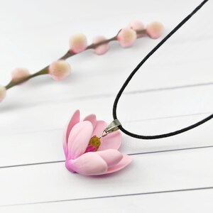 Op de afbeelding: Een delicate roze bloemhangerketting met een zwart koord. De bloem heeft een gedetailleerd ontwerp met een gele kern. Een tak met kleine, roze, donzige knoppen bevindt zich op de achtergrond, alles tegen een witte ondergrond.