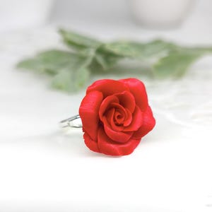 May include: A vibrant red rose ring. The ring features a detailed, three-dimensional rose bloom. The petals are a deep red, and the ring band is silver. The ring is set against a soft, blurred background with green foliage.