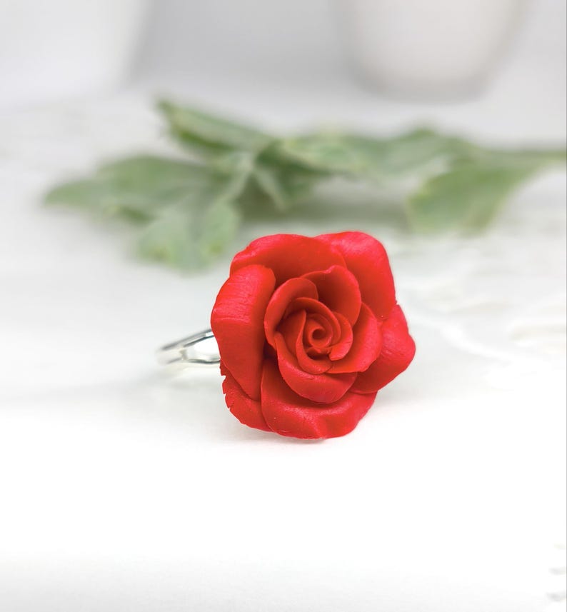 May include: A vibrant red rose ring. The ring features a detailed, three-dimensional rose bloom. The petals are a deep red, and the ring band is silver. The ring is set against a soft, blurred background with green foliage.