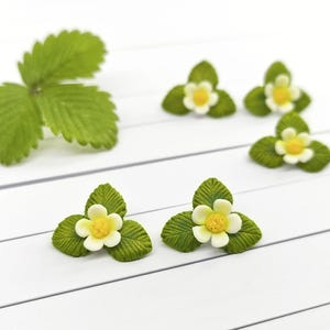 Pode incluir: Close-up de vários pequenos enfeites detalhados de flores e folhas de morango. As flores têm pétalas brancas e centros amarelos, rodeadas por folhas verdes. Uma única folha também é visível. Os itens estão dispostos em uma superfície branca.