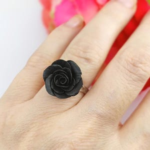 Könnte beinhalten: Ein Ring mit einer mattschwarzen, rosenförmigen Verzierung mit geschichteten Blütenblättern. Die Rose ist auf einem einfachen Metallring befestigt und zeigt ein detailliertes Blumendesign, das auf dem Finger vor einem unscharfen Hintergrund sitzt.
