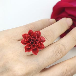Könnte beinhalten: Nahaufnahme eines roten Blumenrings. Der Ring hat ein detailliertes, dreidimensionales Blumendesign mit mehreren Blütenblättern. Der Ring wird an einem Finger getragen, mit einer roten Blume im Hintergrund.