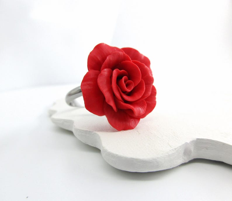 May include: A red rose ring with a detailed floral design. The ring features a large, vibrant red rose as the focal point, set on a silver-colored band. The rose is intricately sculpted, showcasing the delicate petals. The ring is displayed on a white, decorative surface.