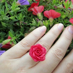 May include: A vibrant red rose ring. The ring features a detailed, three-dimensional rose design. The ring is set against a backdrop of green foliage and red and purple flowers.
