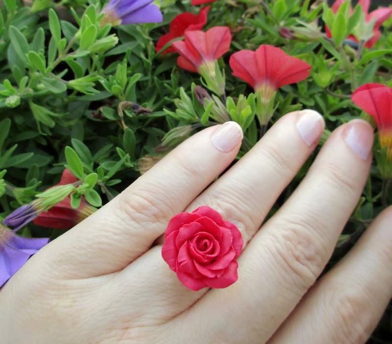 May include: A vibrant red rose ring. The ring features a detailed, three-dimensional rose design. The ring is set against a backdrop of green foliage and red and purple flowers.