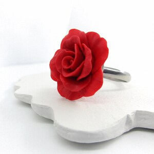 May include: A red rose ring with a detailed floral design. The rose is the focal point, set on a silver-colored band. The ring is displayed on a white, decorative surface.